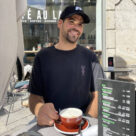Mann sitzt im Straßencafé und hält eine Tasse Cappuccino in der Sonne.
